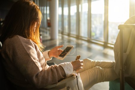 Photo for Side view of anonymous female in casual clothes with luggage browsing smartphone and eating while sitting and waiting for departure in airport room with glass walls in daylight - Royalty Free Image