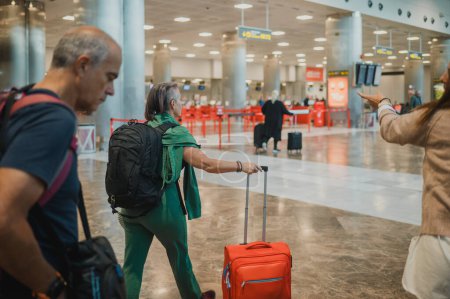 Téléchargez les photos : Sérieux passagers d'avion multiracial en vêtements décontractés avec bagages de voyage marchant dans le hall de départ éclairé de l'aéroport tout en se préparant à monter à bord d'une compagnie aérienne - en image libre de droit