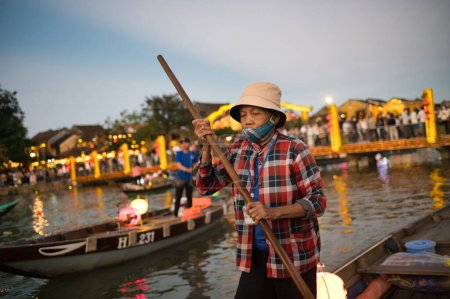 Téléchargez les photos : Calme asiatique mâle en chemise à carreaux et chapeau debout près du bateau avec pagaie tout en flottant dans l'eau de la rivière dans la soirée - en image libre de droit