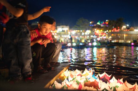 Téléchargez les photos : Plein corps vue latérale de joyeux garçon asiatique en vêtements décontractés souriant et regardant la caméra tout en étant assis sur le bord de la fontaine dans la soirée - en image libre de droit