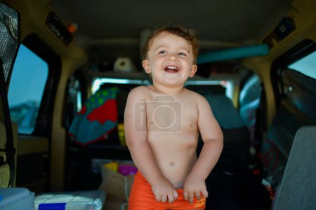 Photo for Happy funny little shitless kid in orange shorts standing inside opened car with many things during road trip - Royalty Free Image
