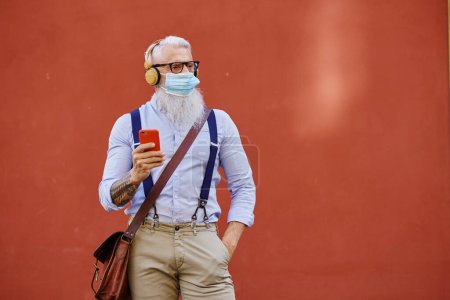 Téléchargez les photos : Homme âgé à la mode avec barbe grise et téléphone portable regardant loin tout en se tenant avec la main dans la poche sur fond rouge - en image libre de droit