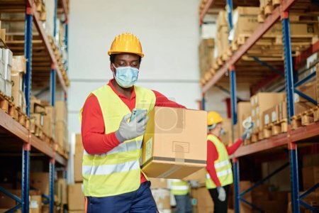 Photo for Low angle of black male employee in workwear and protective mask carrying parcel and checking data on mobile phone while working in logistic service storage during coronavirus pandemic - Royalty Free Image