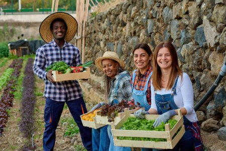 Foto de Cultivadores multiétnicos sonrientes con verduras y frutas variadas en contenedores mirando a la cámara en el campo - Imagen libre de derechos
