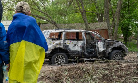Foto de Hombre con bandera ucraniana en el cráter de explosión de drones kamikaze, coche quemado con agujeros de metralla después del mayor ataque de misiles de Rusia contra Ucrania, símbolo de resistencia popular, guerra - Imagen libre de derechos