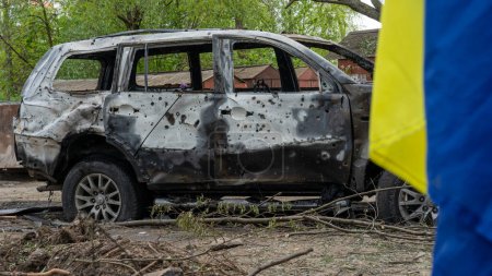 Téléchargez les photos : Drapeau ukrainien devant une voiture incendiée avec des trous d'éclats d'obus après l'explosion d'un missile russe sur l'Ukraine, symbole de la résistance populaire - en image libre de droit