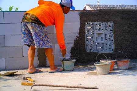 Téléchargez les photos : Bandung, Java occidental, Indonésie - 27 décembre 2023 : Photographie industrielle. Un homme en chemise orange frappe du ciment dans un mur. Plâtrage du mur avec du mortier de ciment. - en image libre de droit