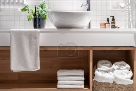 Photo for Wooden countertop with shelf for storage fresh and white terry towels. Wicker laundry basket under washbasin in modern hotel bathroom. Clean textile for body hygiene in comfort apartment - Royalty Free Image