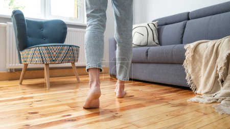 Téléchargez les photos : Plan recadré de jambes féminines marcher pieds nus sur le sol chaud en bois près du canapé dans le salon à la maison, concept de chauffage - en image libre de droit