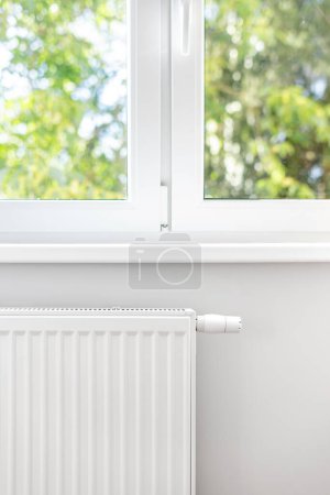 Concept of renovated apartment with new individual heating system installed on wall. Vertical shot of white metal radiator under plastic window frame in bedroom or living room
