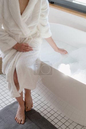 Téléchargez les photos : Vue en angle élevé de la femme portant un peignoir blanc assis au bain, attendant que l'eau chaude remplisse la baignoire. Femme passant la matinée dans la salle de bain. Concept de routine de soins de la peau et procédure de spa à domicile - en image libre de droit