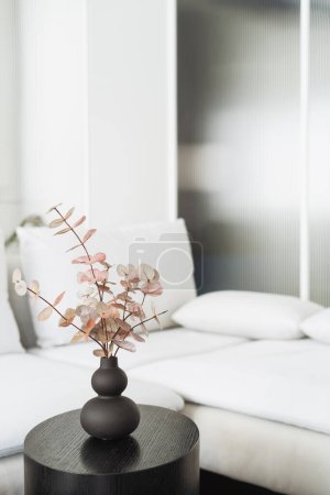 Foto de Rama de eucalipto en jarrón de cerámica sobre mesa auxiliar redonda de madera y sofá con almohadas sobre fondo en apartamento con interior escandinavo - Imagen libre de derechos