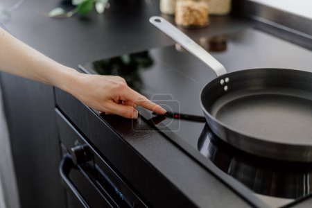 Foto de Botón de sensor táctil de mano femenina en el panel de control de la placa eléctrica y la cena de cocina en la sartén en la cocina casera, electrodomésticos modernos - Imagen libre de derechos