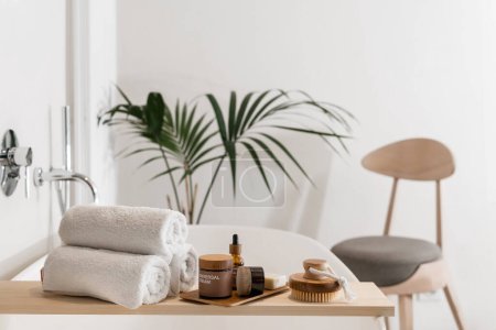 Photo for Skin care cosmetics on bamboo tray, massage peeling brush and fresh terry towels folded on wooden caddy on bathtub, plant and chair in apartment with modern interior - Royalty Free Image