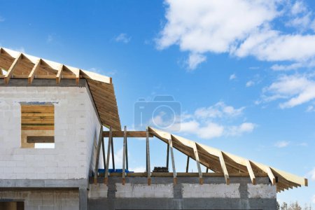 Téléchargez les photos : Vue du bâtiment inachevé depuis les blocs de béton et le toit avec des planches de treillis en bois à l'extérieur sur fond bleu ciel, processus de construction de la maison - en image libre de droit