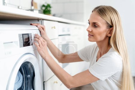 Hausfrau, die das Programm auf dem Bedienfeld der automatischen Waschmaschine auswählt, um die Wäsche im Zimmer in der modernen Wohnung zu waschen. Hauswirtschaftliches Konzept