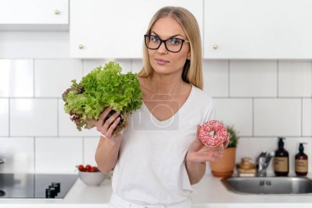 young female standing at home kitchen with green salad leaves and sweet dessert, concept of weight loss diet, choice between healthy and unhealthy food