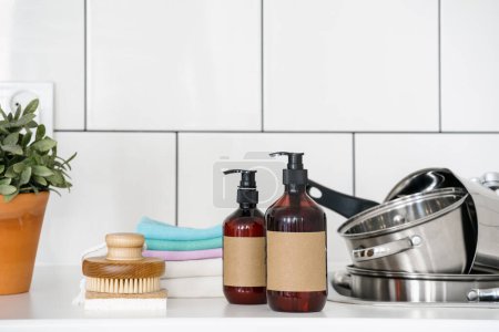 Photo for Front view on soap and dishwashing liquid in dispenser bottles with copy space, eco friendly scrubber sponge and bamboo brush near sink with dirty saucepans at kitchen - Royalty Free Image
