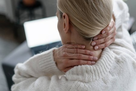 Photo for Back view of female in white sweater feeling discomfort and ache, touching and massaging neck with hands near laptop in room, overwork concept - Royalty Free Image