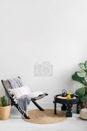 Photo for Lounge rattan chair with cushion and plaid near wooden side table with teapot and cup, houseplants in pots and round floor rug in room with bohemian style - Royalty Free Image