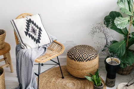 Photo for Angled view on wicker pouf, rattan chair with plaid and pillow, green plants in pots and round rug on floor in stylish living room with ethnic bohemian interior - Royalty Free Image