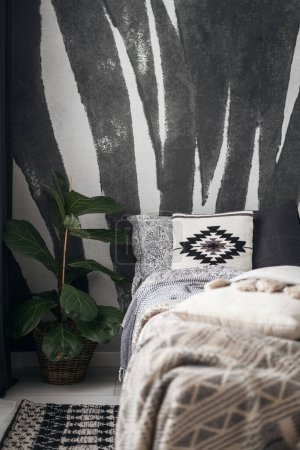 Photo for Detail in bedroom interior with ethnic boho style, comfortable bed with blanket, soft decorative cushions with pattern and potted plant near floor carpet on wall background - Royalty Free Image