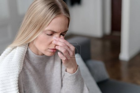 illness female feeling nasal pain or migraine and touching nose, upset girl with fever and grippe wrapped with warm plaid and sitting on sofa at home