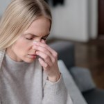 Illness female feeling nasal pain or migraine and touching nose, upset girl with fever and grippe wrapped with warm plaid and sitting on sofa at home