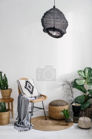 Photo for Home decor in boho style in apartment, rattan chair with plaid and cushion with pattern, wicker pouf near potted plants and round rug on floor on white wall background - Royalty Free Image
