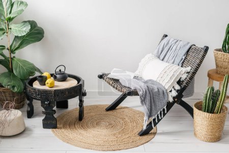 Photo for Cozy interior in room with ethnic boho style, lounge chair with plaid and pillow, wooden coffee table with teapot, cup and fresh fruit, plants in pots and round rug on floor - Royalty Free Image