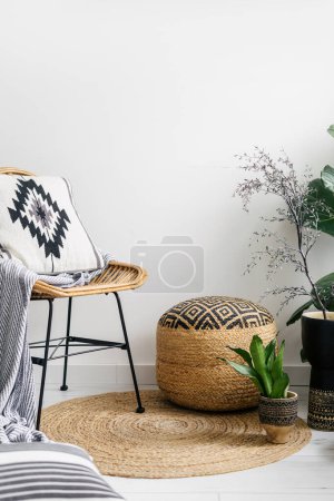 Photo for Detail in apartment with boho interior, rattan chair with soft plaid and cushion in cover with pattern, wicker pouf and potted plants on round rug on floor - Royalty Free Image