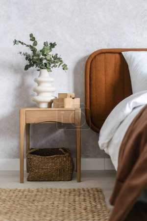 Photo for Detail in bedroom interior, bed with bedclothes, pillow and brown cover, eucalyptus branch in modern vase near bamboo box on wooden side table and wicker basket on floor - Royalty Free Image