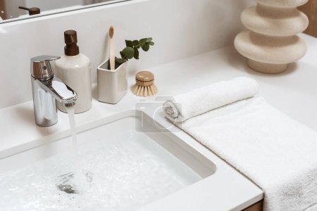 Blickwinkel auf weißes, mit Waschbecken gefülltes Wasser, Metallhahn, Spender, Zahnbürste in einer Tasse, Bambusbürste, gerolltes Handtuch und Keramikvase in einem weißen Möbel