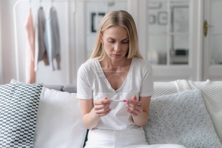 Photo for Confused female looking on positive result at pregnancy test and feeling sad while sitting on sofa in living room, girl thinking about abortion - Royalty Free Image