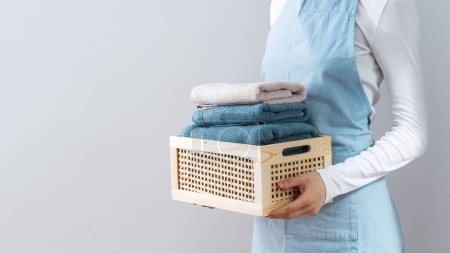 Photo for Cropped view of woman holding wooden wicker basket with folded clean towels against copy space background. Hotel service concept. Female in white shirt and blue apron with fresh textile after laundry - Royalty Free Image