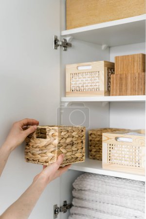 cropped shot of female hands hold wicker basket and put in on shelf in closet with fresh linen and stack of clean towels, organized space concept