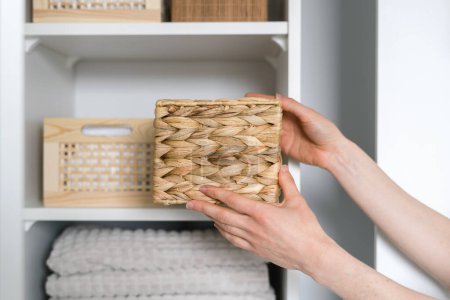 cropped shot of female hands hold wicker storage box and put in on shelf in wardrobe with clothes and clean towels, organized space concept, order inside closet