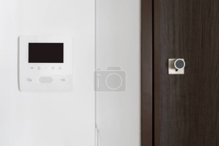 Photo for Close up view of surveillance monitor with electronic doorbell on white wall. Brown wooden door near intercom device with video connection and copy space display. Concept of smart home protection - Royalty Free Image