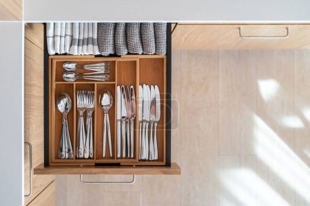 Flat lay view of open kitchen drawer with wooden box for cutlery and folded cotton towels. Separated knives, spoons and forks. Well organized space in the kitchen. Copy space