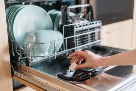 Nahaufnahme einer weiblichen Hand, die ein Geschirrspüler-Tablet in eine offene Einbauspülmaschine mit Geschirr in der modernen Wohnküche einsetzt. Haushalt, Hauswirtschaft, häusliches Konzept
