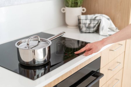 cropped shot of female hand turn on induction stove and choose temperature for cooking dinner in stainless steel saucepan with lid at kitchen, modern household appliances concept