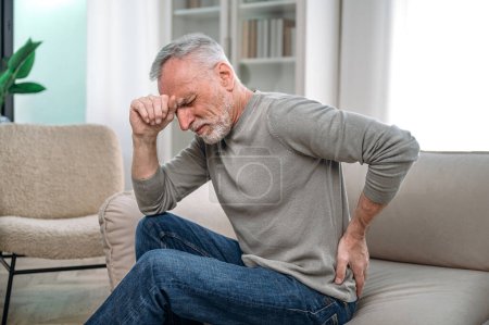 Old mature man sitting on couch, feeling sudden back pain and suffering from ache. Rheumatism and low-back lumbar illness concept. Waiting for visit in clinic