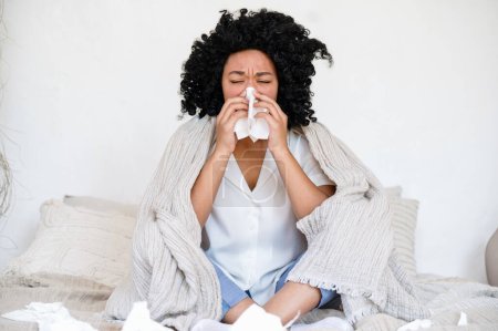 Photo for Sick african american woman sitting on bed covered with blanket, suffering from viral disease and blowing nose in paper tissue. ill female has seasonal allergy - Royalty Free Image