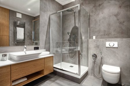 Badezimmer mit luxuriösem Interieur, Glasduschkabine, WC-Schüssel mit verstecktem Zisternen-Tank, Keramik-Waschbecken mit Wasserhahn und Holzunterschrank mit sauberem Handtuch