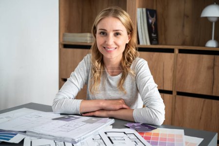 Portrait of smiling young interior designer at home working place. Pantone swatches and house architecture construction plans on table. Choosing best colors for renovating