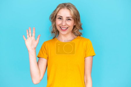 Téléchargez les photos : Portrait de jeune femme souriante regardant la caméra et agitant la main isolée sur fond bleu studio. Salutation, bonjour ou au revoir. Concept de communication. - en image libre de droit
