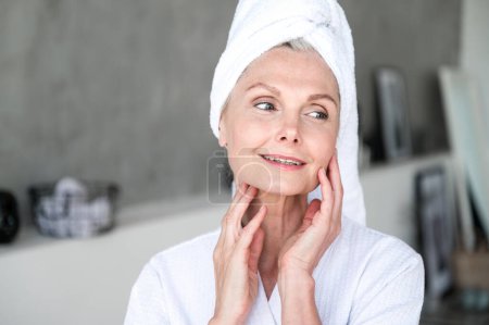 Mujer sonriente de 50 años con toalla en la cabeza mirando hacia otro lado, tocando piel sensible suave hidratada y saludable en la mejilla y el cuello, haciendo hidratación matutina, tratamiento de belleza spa. Concepto de cuidado facial