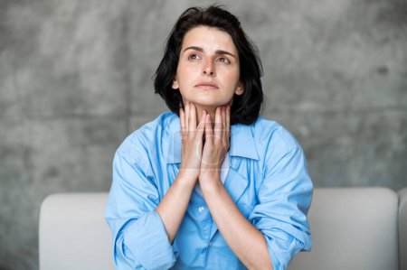 Young upset woman with throat sore, feeling unwell and touching her neck while sitting on sofa. Sick female with inflamed lymph nodes and flu symptoms. Healthcare concept