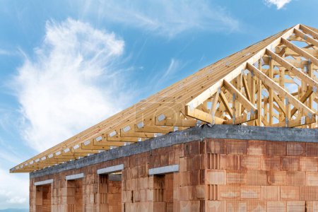 Dach mit Holzbalken über Ziegelwänden. Beispiel für ein neues Haus im Bau. Teil des unfertigen Hauses mit Holzdach vor blauem Himmel und Kopierfläche für Werbekonzepte