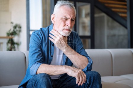 Middle aged man suffering sore throat, touching neck and feeling sick while sitting alone on couch in living room. Unhappy frowning male has viral, infection or flu symptoms